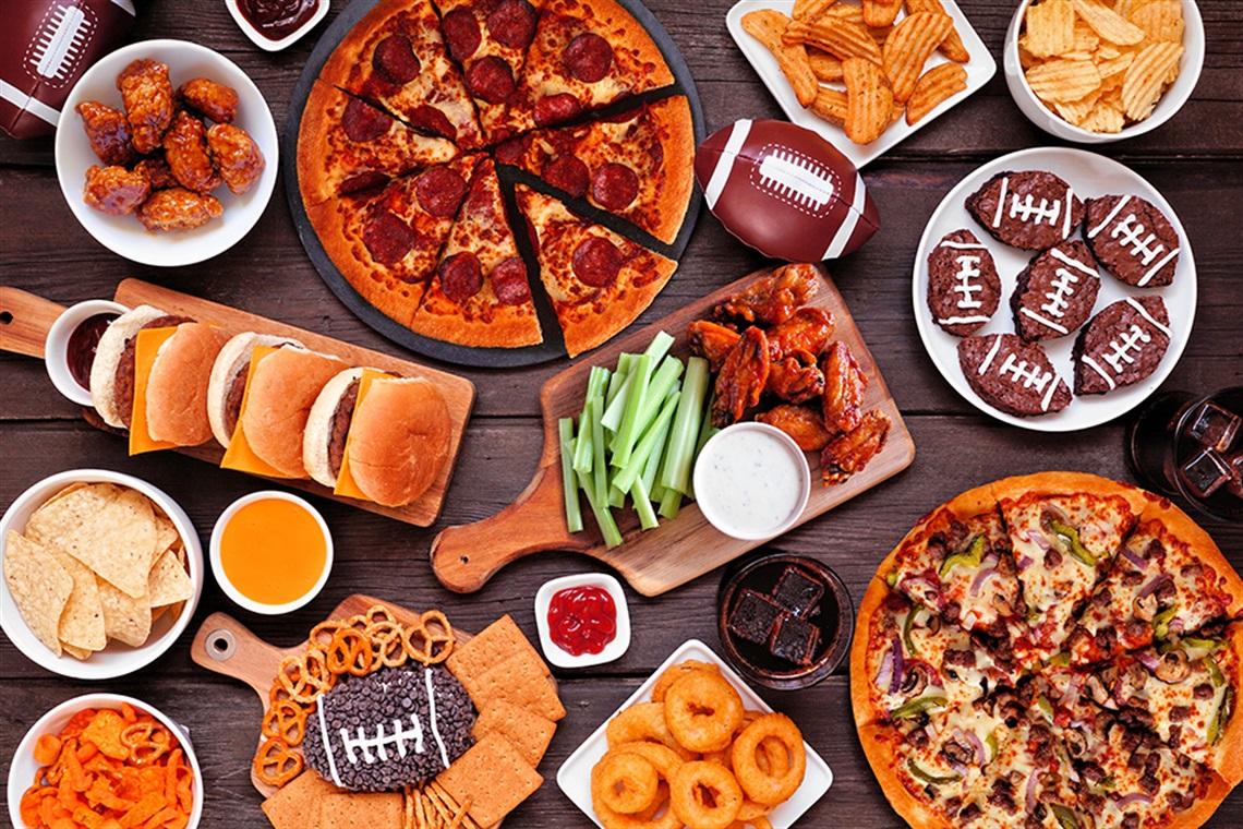 Football theme food table scene. Pizza, hamburgers, wings, snacks and sides. Overhead view on a dark wood background.