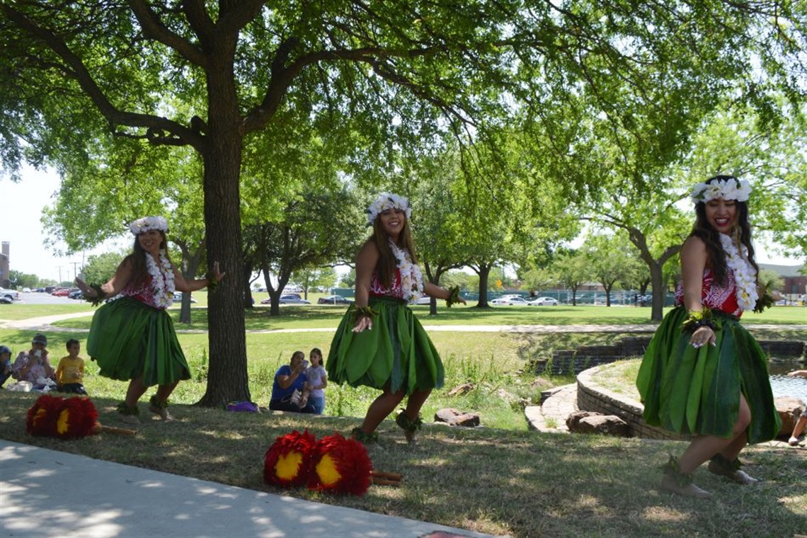 Three dancers in traditional Hawaiian attire are performing under a tree beside a park lawn.