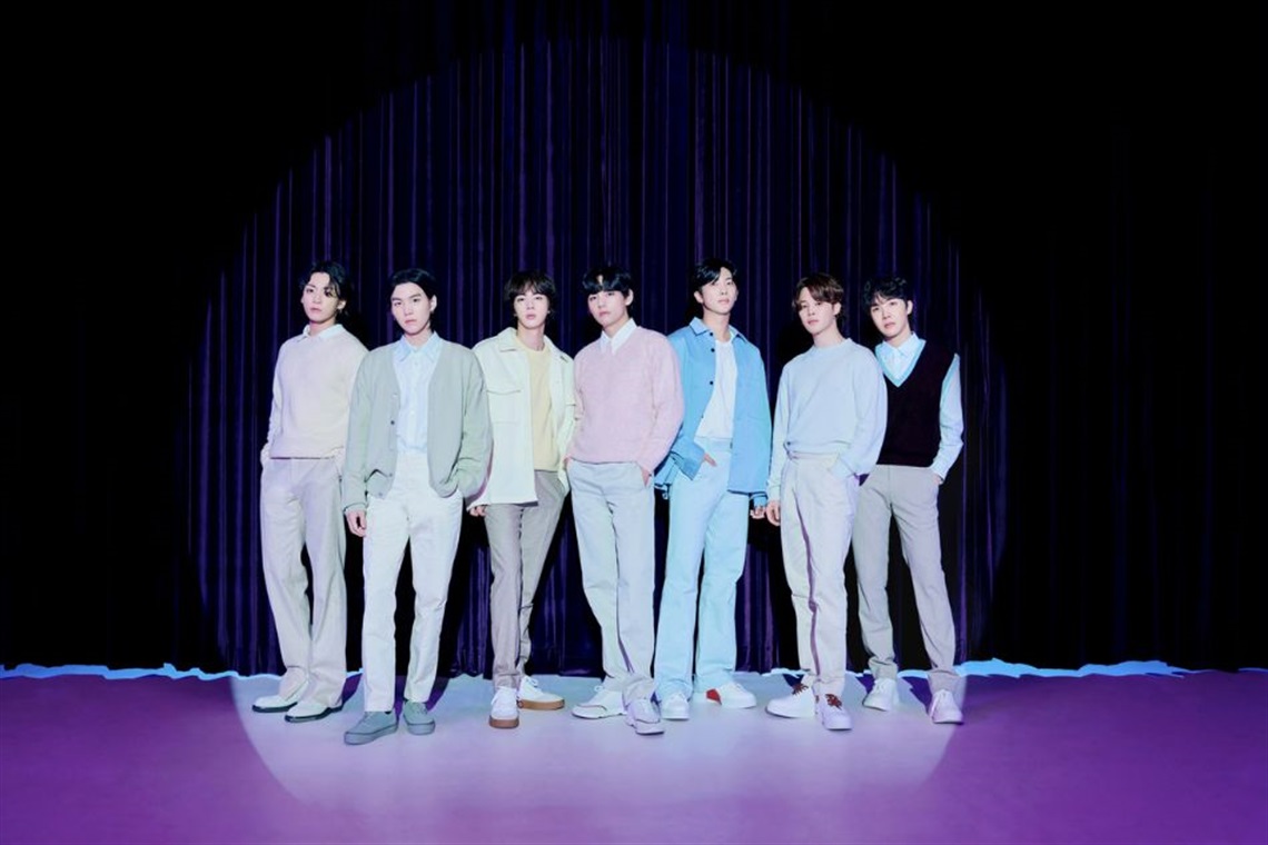 Group photo of BTS standing in a row on a stage with a purple background, dressed in pastel-colored outfits.