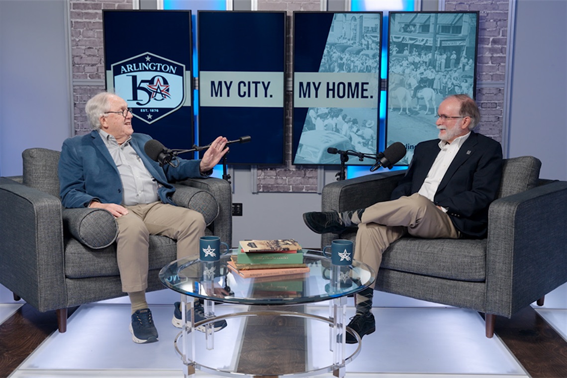 OK Carter, left, speaks as Gerald Saxon, right, laughs. Both are seated. Background image on TV screens say 