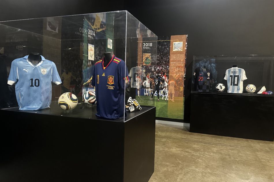 A display case containing two sports jerseys, a soccer ball, a shoe, and a small display screen.