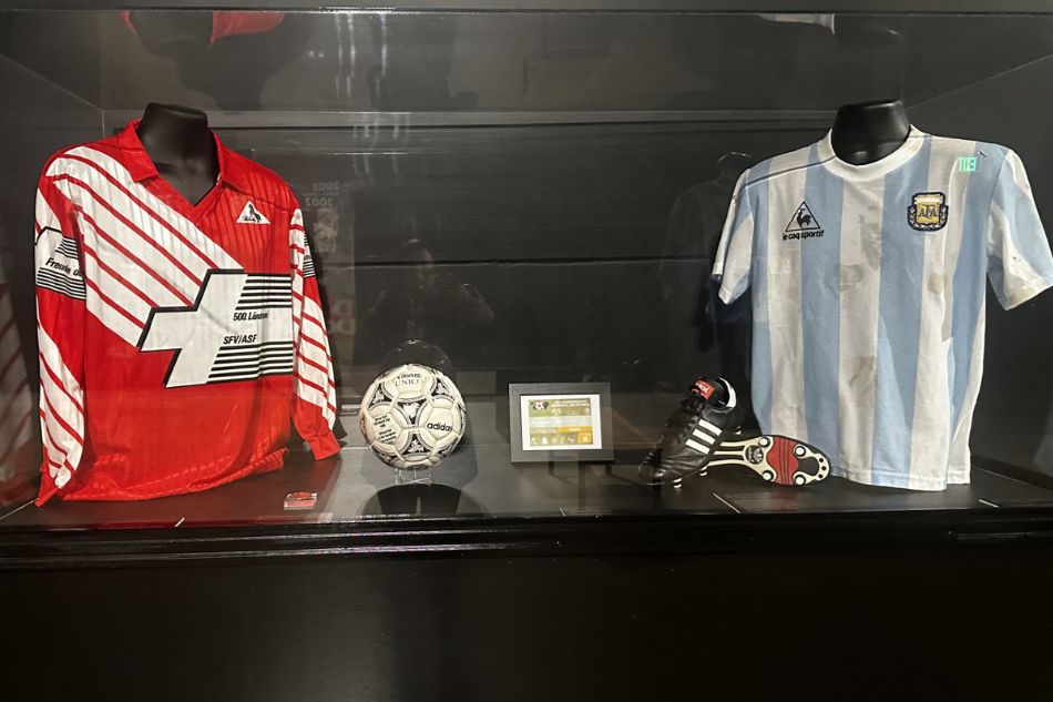 A display case containing two sports jerseys, a soccer ball, a shoe, and a small display screen.