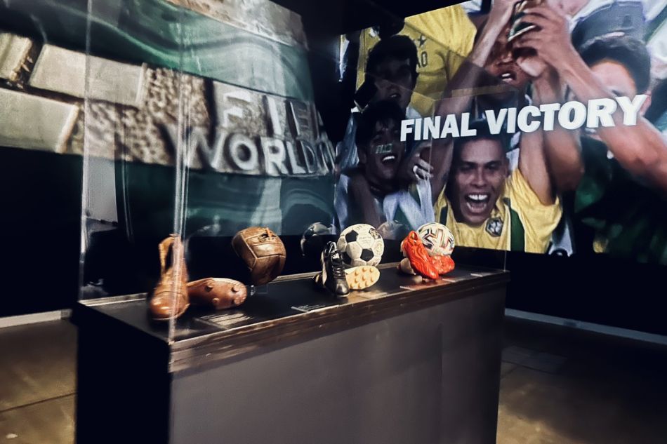 A display case features vintage soccer balls and a shoe. In the background, there's a large photo of cheering fans wearing yellow jerseys, with the words 