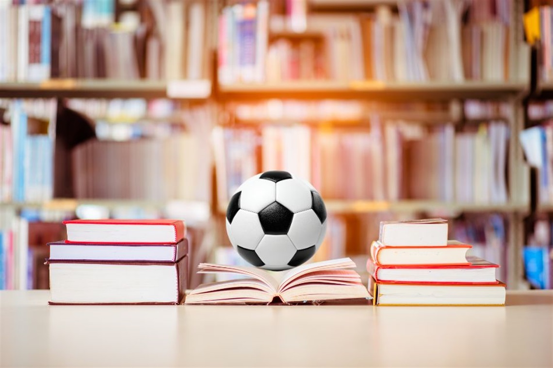 A soccer ball resting on an open book, surrounded by stacks of books