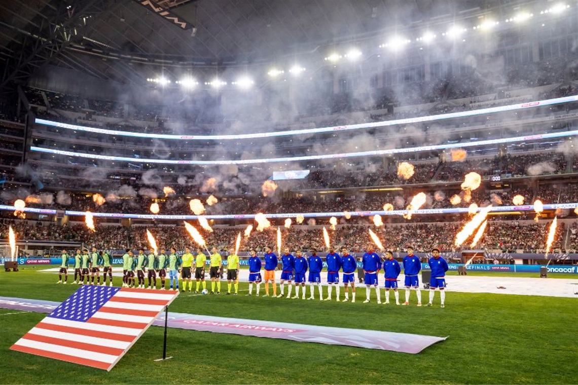 Teams line up on the field for a soccer match, with a large American flag displayed in the foreground and fireworks above