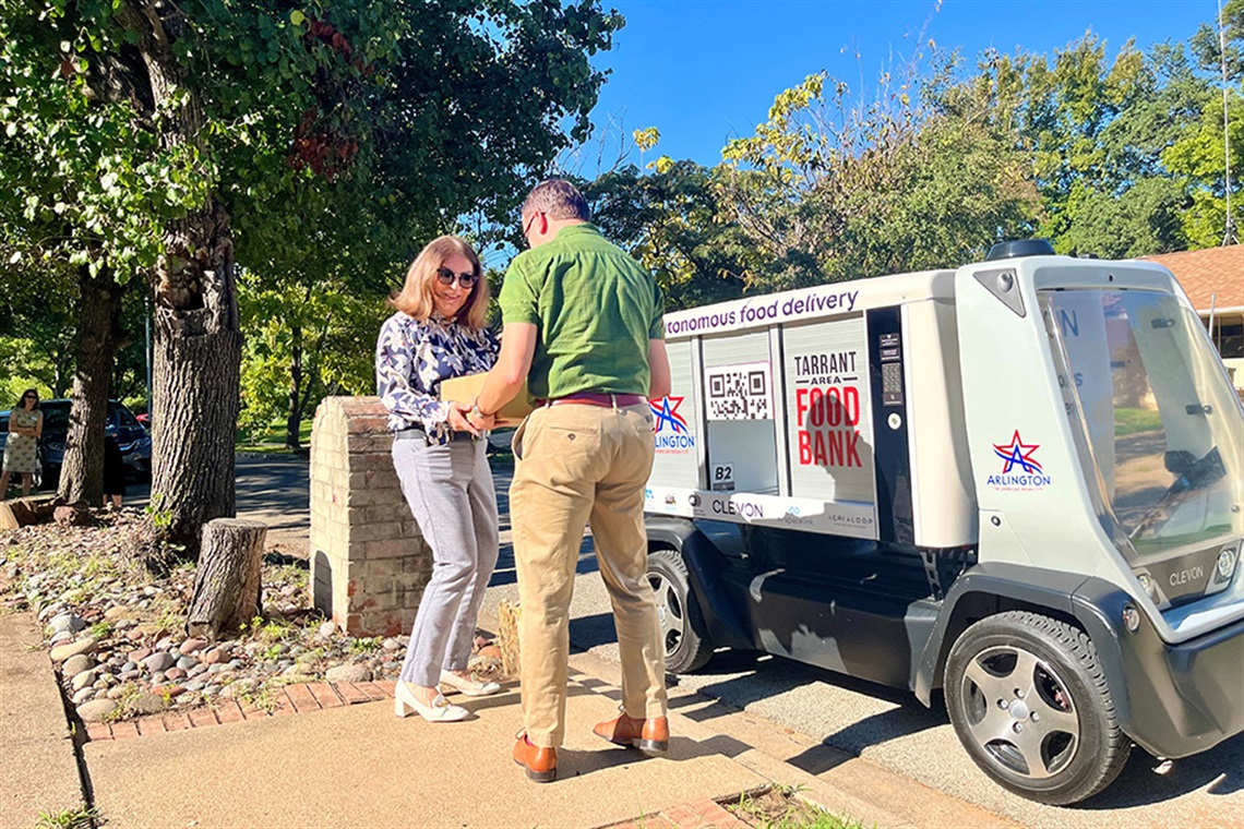 A man, standing on the sidewalk in front of a four-wheeled robot with a cargo bay, hands a box of donated food to a woman in front of a home.