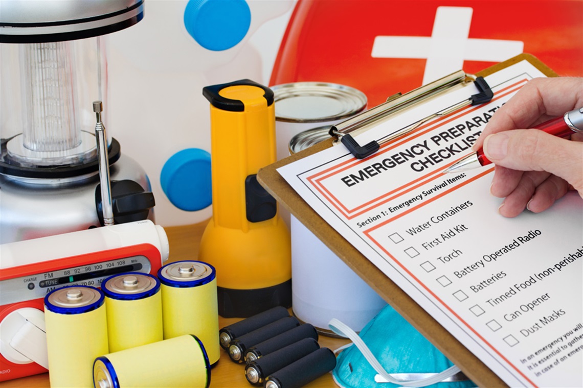 Pen hovering over Emergency Preparedness checklist with emergency supplies in the background